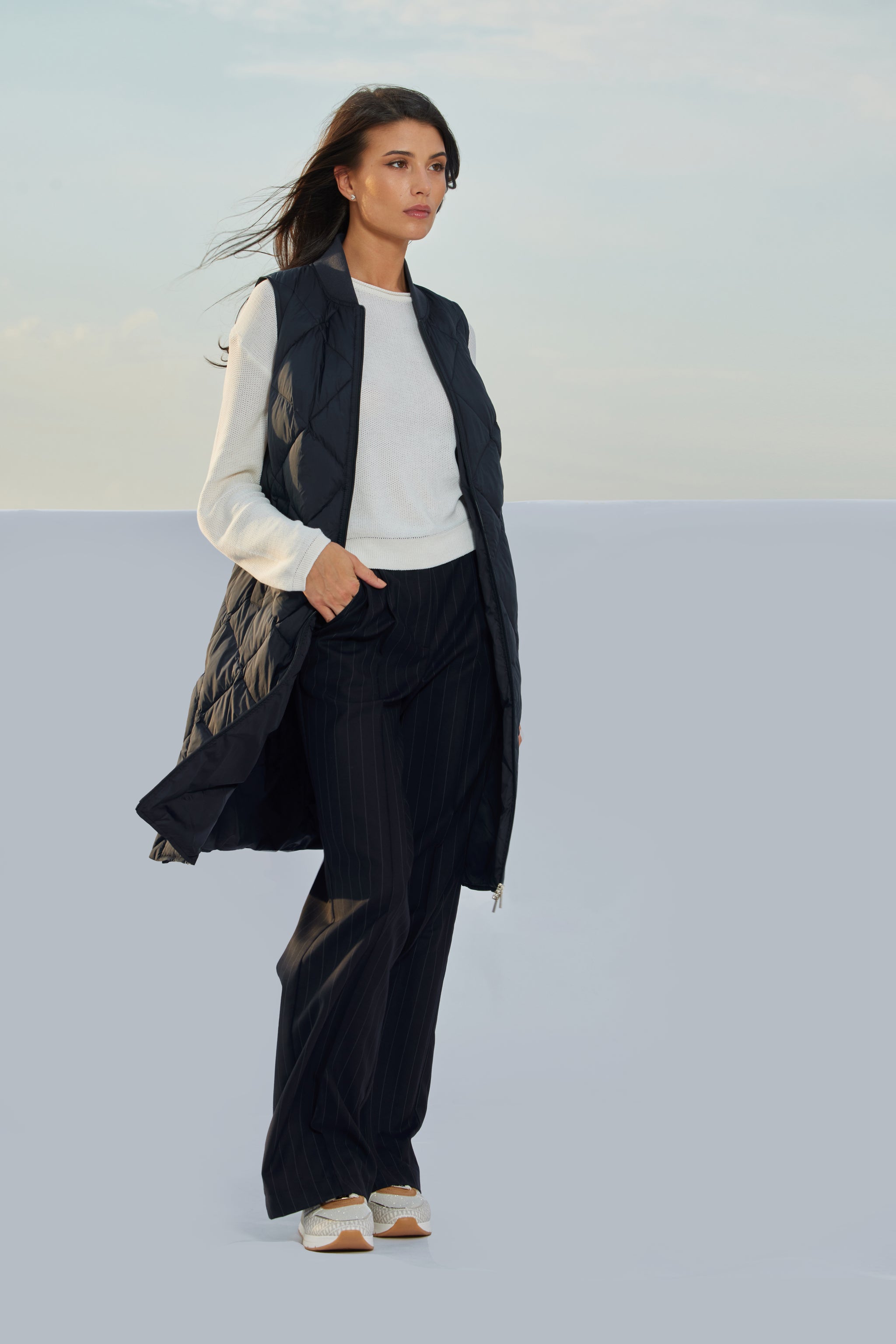 Vestă dark navy damă Madzerini, textură matlasată, purtată cu bluză albă și pantaloni în dungi negri.